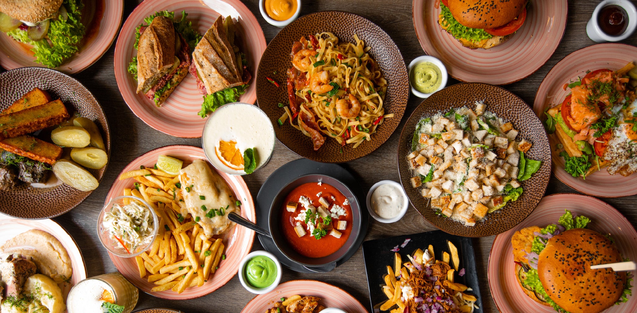 Various plates of food on a wooden table, including sandwiches, french fries, soups, salads, sauces, and drinks.